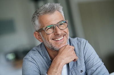 Man with gray hair in gray shirt in brown glasses smiling leaning his chin on his hand